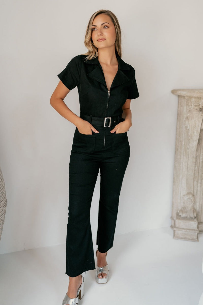Full body view of model wearing the Rielle Black Adjustable Belt Short Sleeve Jumpsuit which features black fabric, two front pockets, a front zipper, an adjustable belt with a silver buckle, a collared neckline, short sleeves, and straight pant legs.