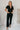 Full body view of model wearing the Rielle Black Adjustable Belt Short Sleeve Jumpsuit which features black fabric, two front pockets, a front zipper, an adjustable belt with a silver buckle, a collared neckline, short sleeves, and straight pant legs.