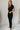 Full body side view of model wearing the Rielle Black Adjustable Belt Short Sleeve Jumpsuit which features black fabric, two front pockets, a front zipper, an adjustable belt with a silver buckle, a collared neckline, short sleeves, and straight pant legs