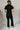 Full body back view of model wearing the Rielle Black Adjustable Belt Short Sleeve Jumpsuit which features black fabric, two front pockets, a front zipper, an adjustable belt with a silver buckle, a collared neckline, short sleeves, and straight pant legs