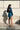 Back view of model wearing The Elaine Satin Dress features a blue, green satin material, a cowl neckline, long sleeves, an open cowl back with a tie closure, and a mini length.