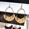 Front view of the Flower Power Earrings in mustard, which feature wire arches with crescent pendants with mustard, orange, and green flower print.