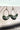 Front view of the Flower Power Earrings in sage, which feature white arches with crescent pendants, with a green floral pattern.