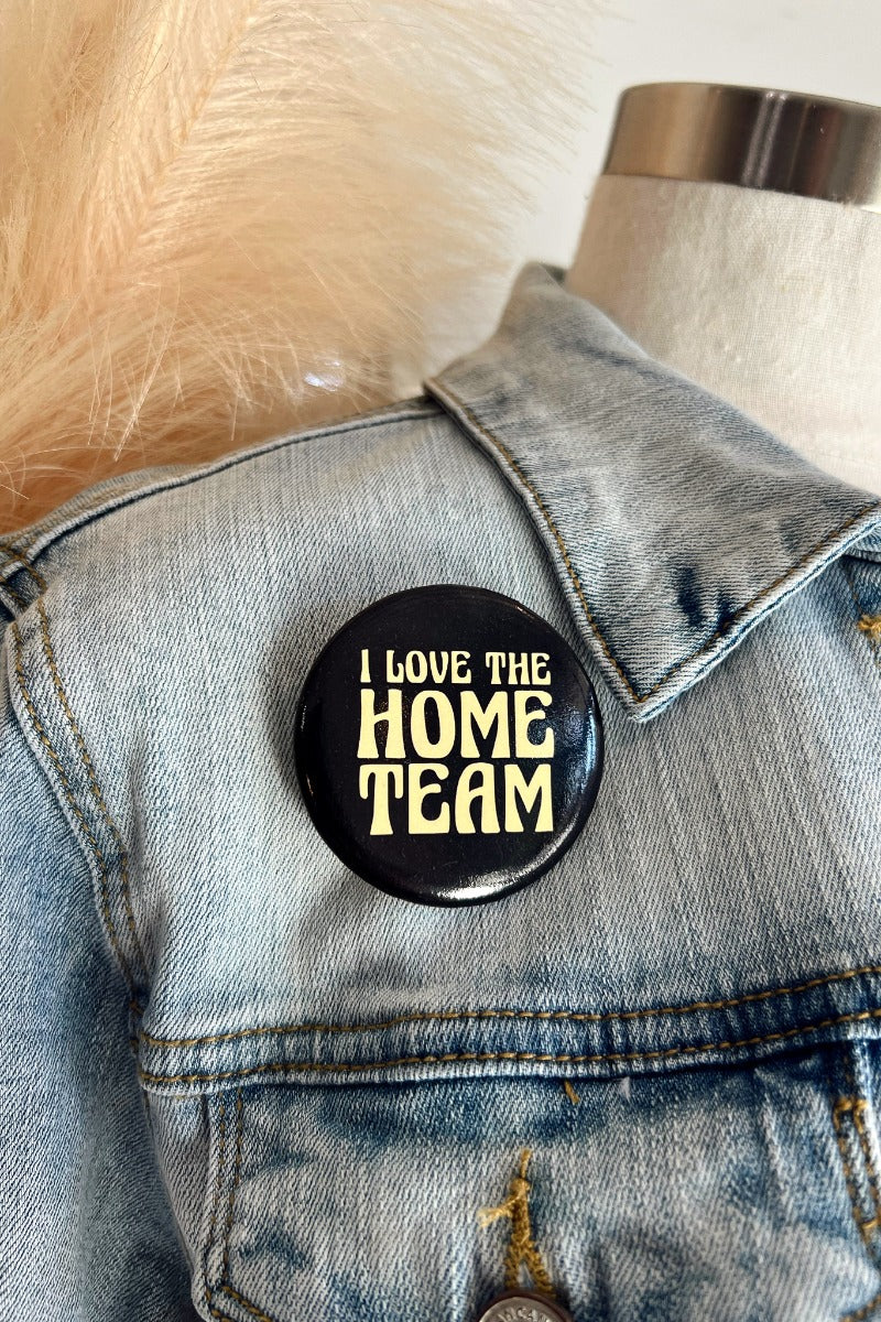 Close up image of the I Love The Home Team Button, that features a black background with yellow text. Button is pinned on denim jacket.