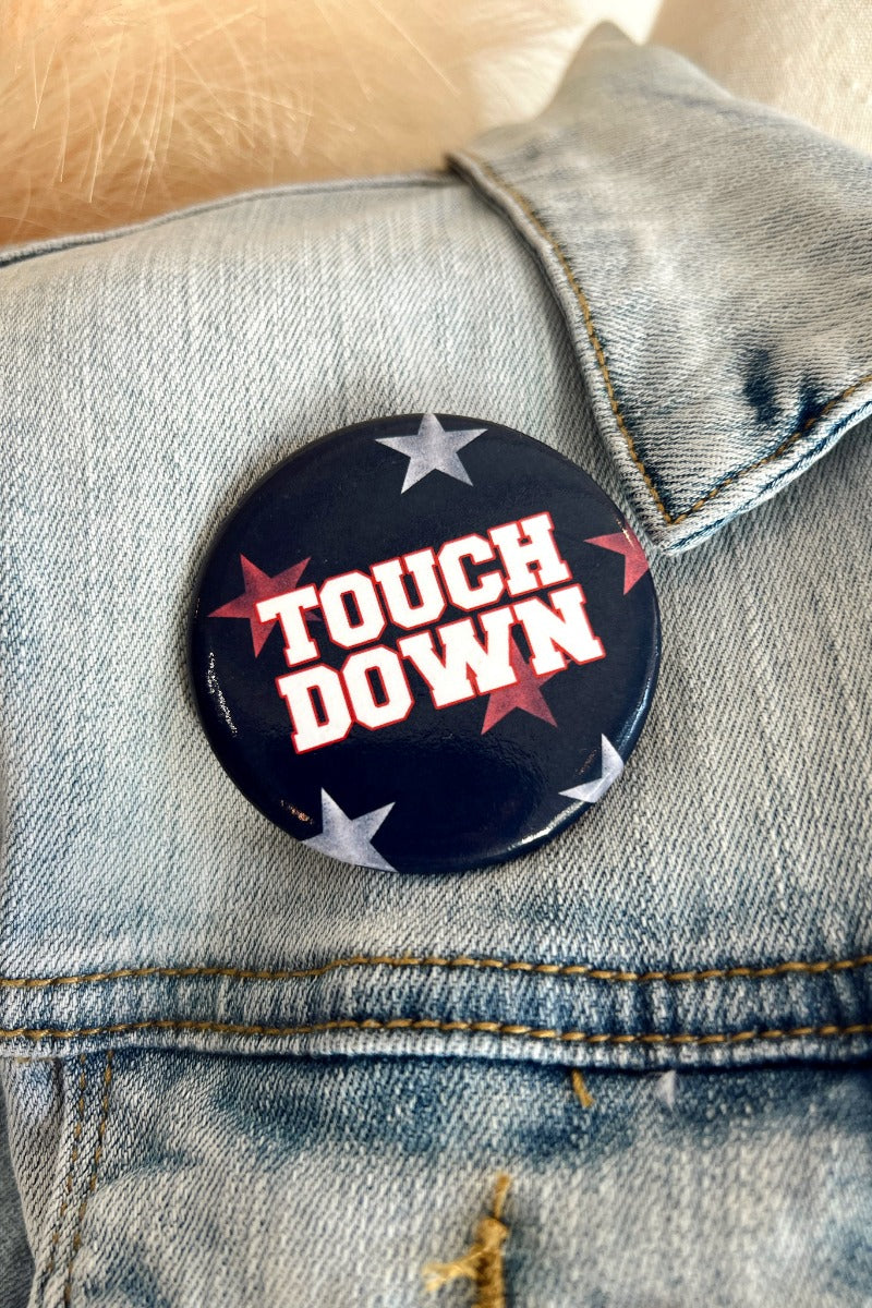 Close-up image of the Touch Down Button, that features a navy button with grey and red stars. Text says "TOUCH DOWN" in white. Button shown on denim jacket.