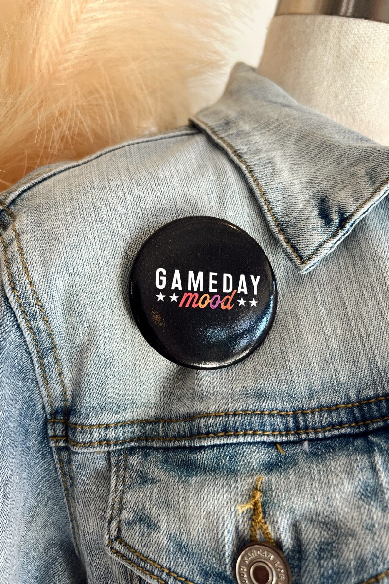 Close up image of the Gameday Mood Button which features a black button with white stars. Text says "GAMEDAY" in white and "Mood" in multicolor. Button is pinned to a denim jacket.