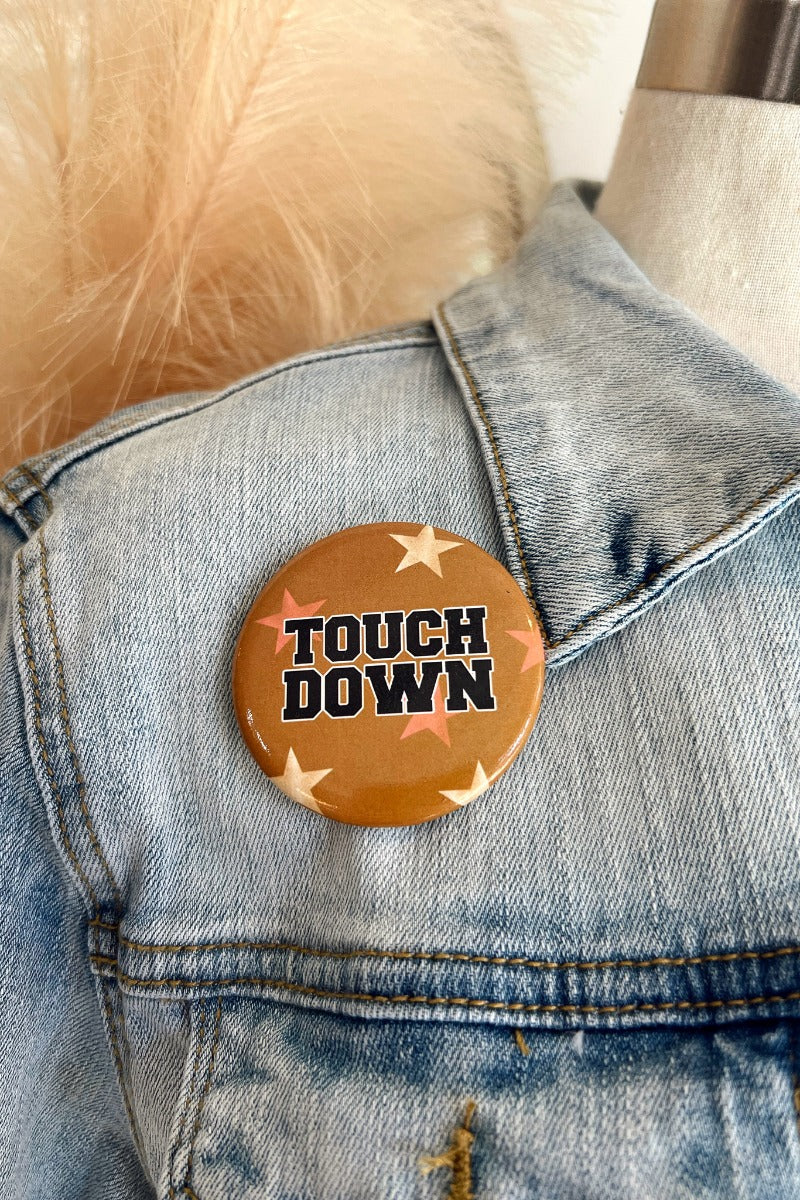 Close up image of the Touch Down Button in Gold features a mustard button with beige and orange. Text says "TOUCH DOWN" in black. Button is pinned to a denim jacket.