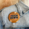 Close up image of the Touch Down Button in Gold features a mustard button with beige and orange. Text says "TOUCH DOWN" in black. Button is pinned to a denim jacket.