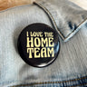 Close up image of the I Love The Home Team Button, that features a black background with yellow text. Button is pinned on denim jacket.