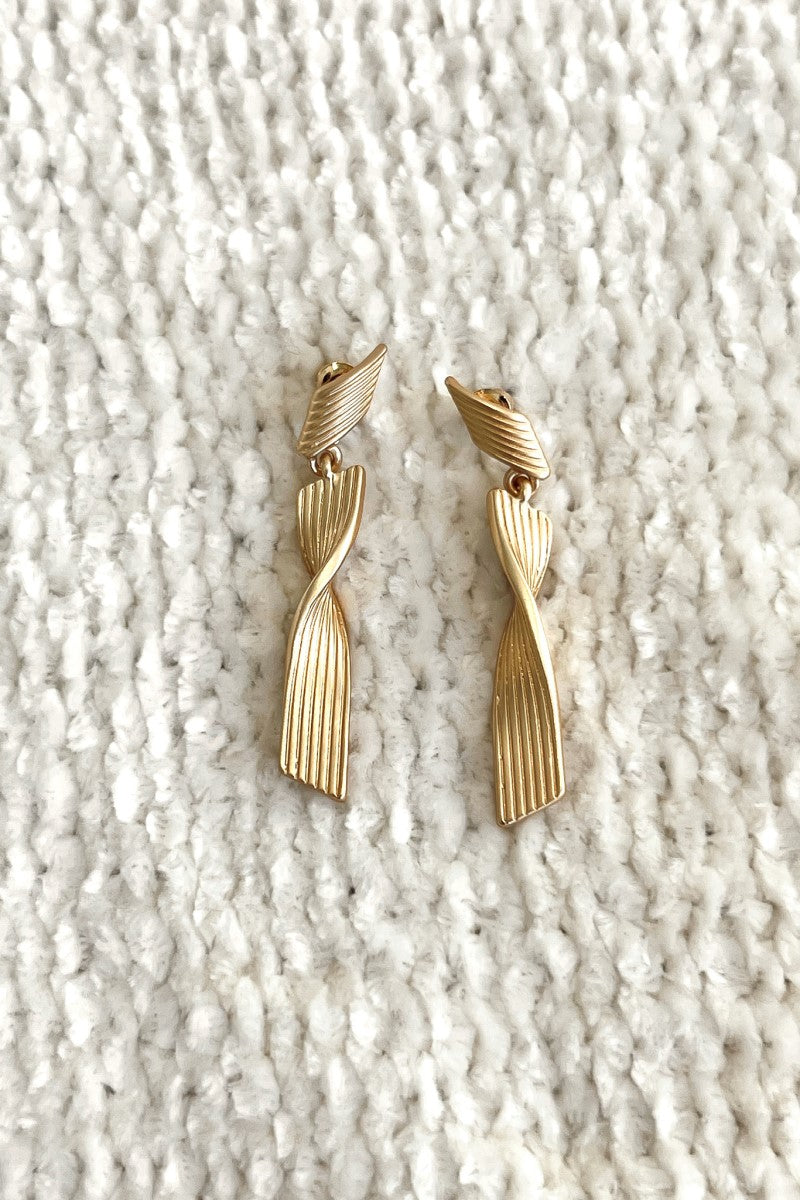 Close up image of the All Twisted Up Earrings that feature matte gold swirl dangles with pleated details.