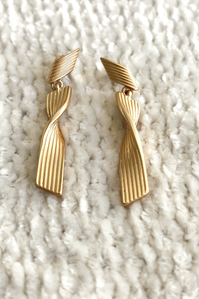 Close up image of the All Twisted Up Earrings that feature matte gold swirl dangles with pleated details.