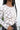 Close-up front view of model wearing the New Rhythm Top, that has cream and beige fabric with a dimensional pattern, ruching ties on the hem, a round neckline and long sleeves.