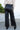 Back view of model wearing the Center of Attention Pants, that feature black faux leather fabric, two front pockets, a zipper and button closure, belt loops, and wide legs