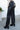 Side view of model wearing the Center of Attention Pants, that feature black faux leather fabric, two front pockets, a zipper and button closure, belt loops, and wide legs
