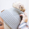 Close side view of model wearing the Anchorage Beanie in Grey, that features grey knit fabric with a stitched pattern, a front patch that says "C.C exclusives", and a brown faux-fur pompom.
