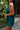 Side view of model wearing the Skylar Satin Dress in Teal which features teal satin fabric, halter neckline with a tie, ruching on the sides, back zipper with hook closures and lining.