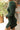 Side view of model wearing the Unforgettable Dress which features green sheen fabric, smalls slits on each side of the hem, ruching on the sides, round neckline, lining and long sleeves.