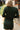 Close up back view of model wearing the Unforgettable Dress which features green sheen fabric, smalls slits on each side of the hem, ruching on the sides, round neckline, lining and long sleeves.