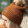 Side view of model wearing the Anchorage Beanie in brown, that has brown knit fabric with a stitched pattern, a front patch that says "C.C exclusives", and a brown faux-fur pompom.
