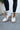 Close-up view of model wearing the  Brighton Lace-Up Boot, that has an off white faux-leather and nylon upper, a taupe rubber sole with a 3" heel, and cognac laces.