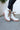 Close-up side view of model wearing the  Brighton Lace-Up Boot, that has an off white faux-leather and nylon upper, a taupe rubber sole with a 3" heel, and cognac laces.