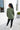 Full body back view of model wearing the This Is Easy Top in Olive which features green fabric, hem detailing, a high-low hem with slits, a round neckline, and dropped shoulder balloon long sleeves.