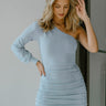 Close up view of model wearing the Making New Rules Dress which features light blue sheen fabric, one shoulder with a long sleeve, lining and ruched detailing along the sides.