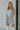 Close up view of model wearing the Making New Rules Dress which features light blue sheen fabric, one shoulder with a long sleeve, lining and ruched detailing along the sides.