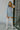Full body view of model wearing the Making New Rules Dress which features light blue sheen fabric, one shoulder with a long sleeve, lining and ruched detailing along the sides.