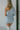Back view of model wearing the Making New Rules Dress which features light blue sheen fabric, one shoulder with a long sleeve, lining and ruched detailing along the sides.
