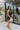 Side view of the City Walk Dress that features a black silky material, a cowl neckline, a sleeveless design with thin adjustable straps, a tie at the waist, a draped design, a back zipper closure, and a mini length.