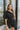 Frontal view of the Juniper Romper that features a black material, an asymmetrical neckline, a one shoulder short puffy sleeve, pockets, a waist tie detail, and a hidden side zipper.