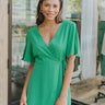 Frontal view of the Speechless Romper that features a green chiffon material, a surplice neckline, a short sleeve with a slit, a waist tie detail, a faux wrap skirt overlay, a shorts back, and a back zipper closure.