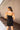 Back view of model wearing the Show Stopper Dress in Black, which features a black chiffon material, a straight neckline, a sleeveless design with thin straps, and a ruched overlapped detail on the skirt