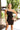 Front view of model wearing the Always A Classic Dress, which features a black chiffon material, a straight neckline, a sleeveless design with a one shoulder thin strap, a twisted knot detail at the waist, and a mini length