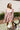 Front view of model wearing the Lost In A Daydream Dress, which features a blush colored material, a white and orange floral print, a V neckline, a long sheer sleeve with smocked detailing at the wrist, a ruffle detail at the bottom