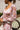 Close-up view of model wearing the Lost In A Daydream Dress, which features a blush colored material, a white and orange floral print, a V neckline, a long sheer sleeve with smocked detailing at the wrist, a ruffle detail at the bottom