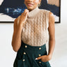 Front view of model wearing the Meet Me In Maine Sweater Tank in Taupe that has a stretchy cable knit material, a turtleneck, a sleeveless design, and a thick bottom hem. Tucked into shorts.
