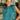 Front view of model wearing the En Vogue Bodysuit that has a textured green material with sheen, a high neck with a tie back, a ruched sleeveless design with shoulder pads, an open back, a bikini bottom, and a snap closure. Worn with white jeans.