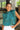 Front view of model wearing the En Vogue Bodysuit that has a textured green material with sheen, a high neck with a tie back, a ruched sleeveless design with shoulder pads, an open back, a bikini bottom, and a snap closure. Worn with white jeans.