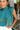 Close-up front view of model wearing the En Vogue Bodysuit that has a textured green material with sheen, a high neck with a tie back, a ruched sleeveless design with shoulder pads, an open back, a bikini bottom, and a snap closure. Worn with white jeans.