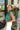 Side view of model wearing the En Vogue Bodysuit that has a textured green material with sheen, a high neck with a tie back, a ruched sleeveless design with shoulder pads, an open back, a bikini bottom, and a snap closure. Worn with white jeans.