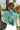 Back view of model wearing the En Vogue Bodysuit that has a textured green material with sheen, a high neck with a tie back, a ruched sleeveless design with shoulder pads, an open back, a bikini bottom, and a snap closure. Worn with white jeans.