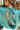 Close-up back view of model wearing the En Vogue Bodysuit that has a textured green material with sheen, a high neck with a tie back, a ruched sleeveless design with shoulder pads, an open back, a bikini bottom, and a snap closure. Worn with white jeans.