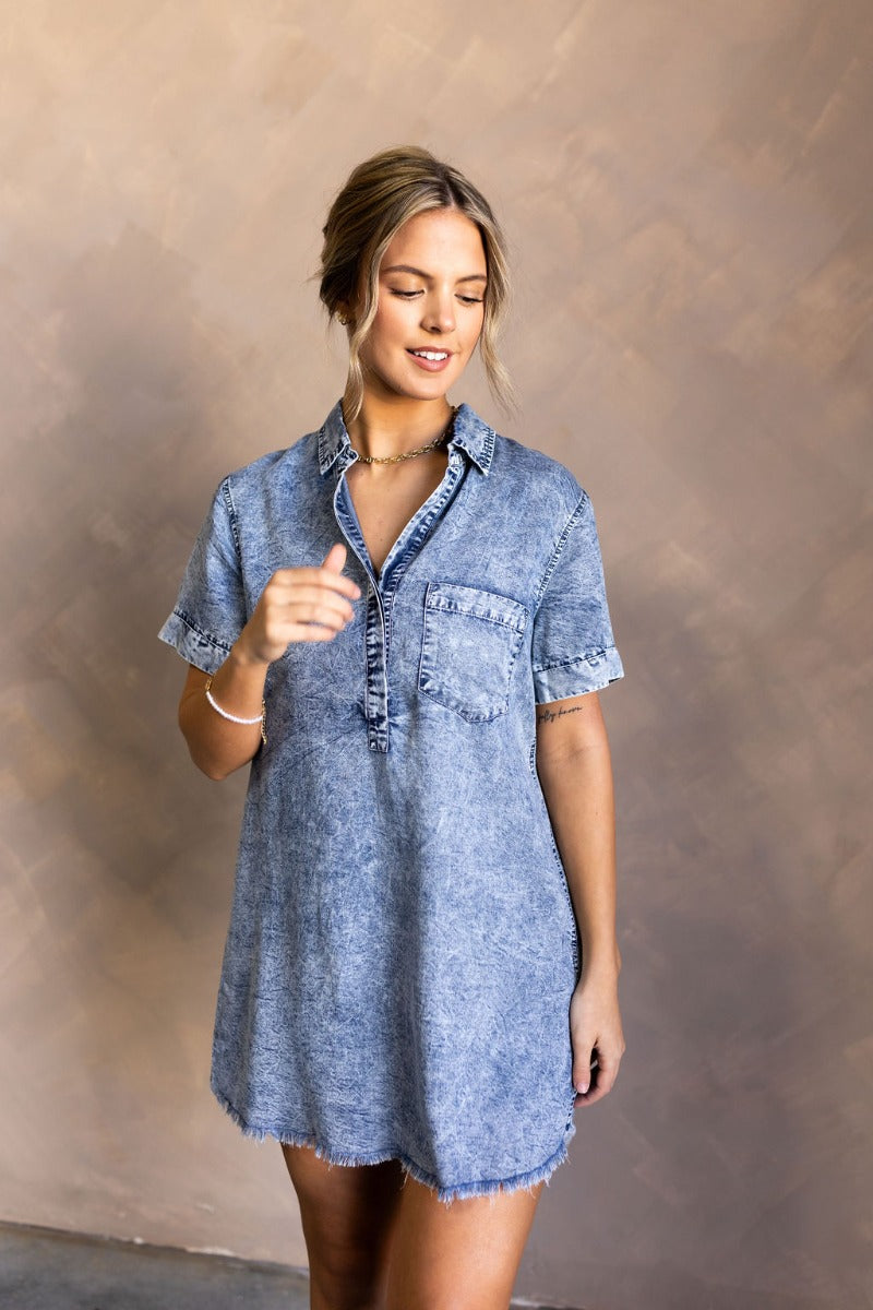 front view of model wearing the Aubrie Washed Blue Short Sleeve Mini Dress that has washed light blue chambray fabric, a chest pocket, a hidden quarter button-up, a v neck, a collar, a frayed hem, and cuffed short sleeves.