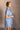 Side view of model wearing the Aubrie Washed Blue Short Sleeve Mini Dress that has washed light blue chambray fabric, a chest pocket, a hidden quarter button-up, a v neck, a collar, a frayed hem, and cuffed short sleeves.