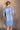 Back view of model wearing the Aubrie Washed Blue Short Sleeve Mini Dress that has washed light blue chambray fabric, a chest pocket, a hidden quarter button-up, a v neck, a collar, a frayed hem, and cuffed short sleeves.