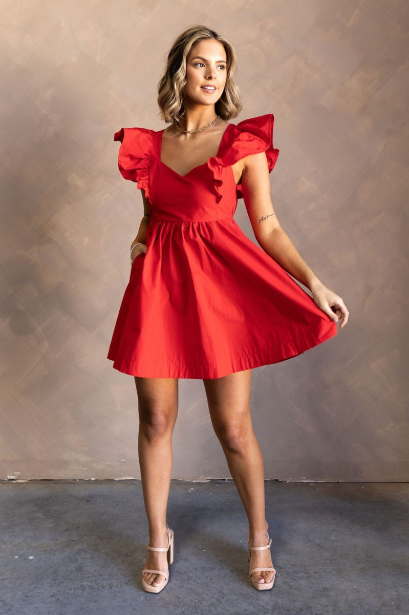Full body front view of model wearing the Ava Red Ruffle Bow-Back Mini Dress that has red cotton fabric, mini length, flare skirt, pockets on each side, a sweetheart neck, ruffle straps, and an open back with tie closures.