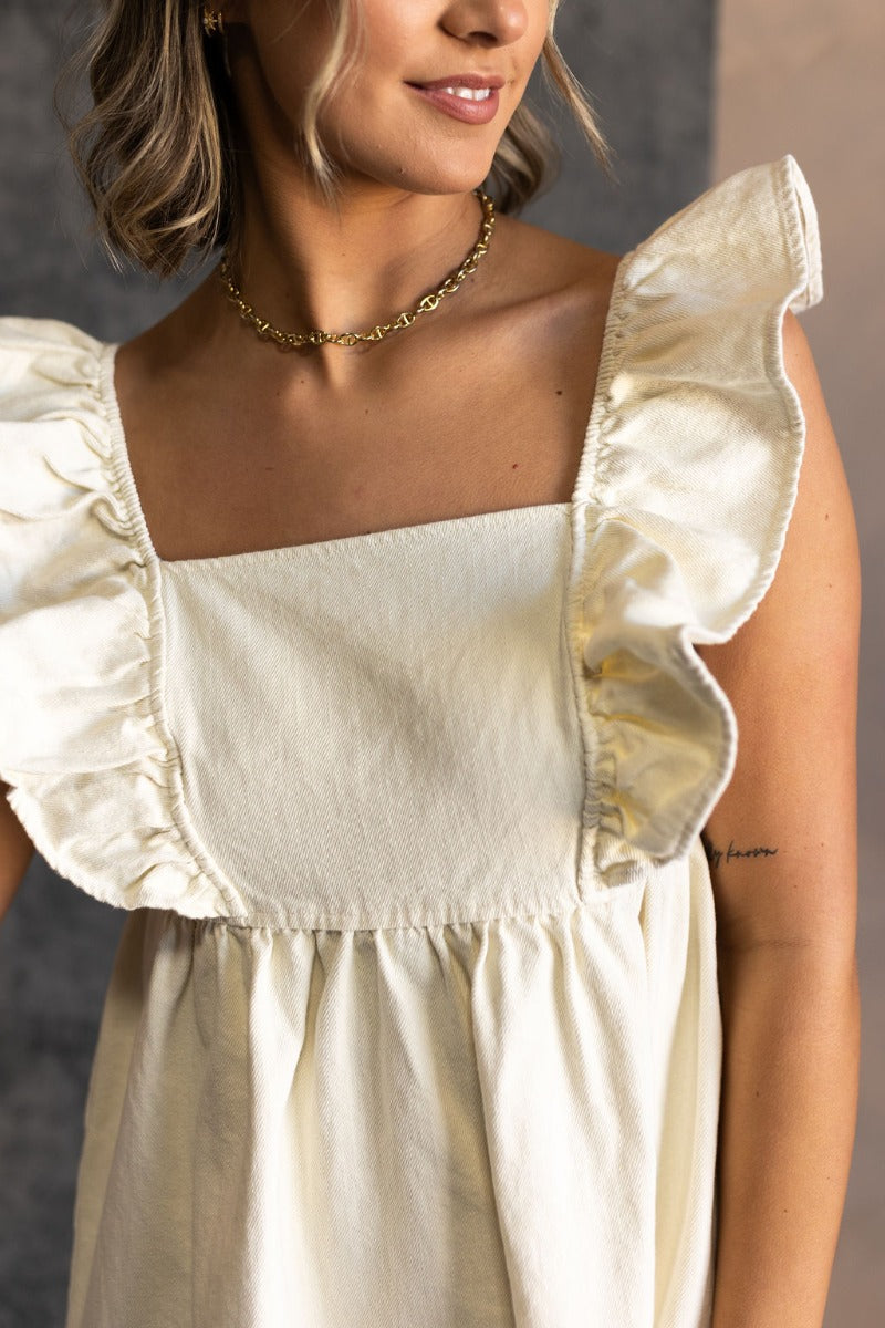 Close upper front view of model wearing the Rory Cream Ruffle Sleeveless Mini Dress that features cream cotton denim fabric, mini length, a square neckline, ruffle straps, and a smocked back.