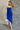 Full body view of model wearing the Liliana Blue Asymmetrical Sleeveless Midi Dress which features royal blue knit fabric, an asymetrical ruffle hem, midi length, a square neckline, and adjustable straps.
